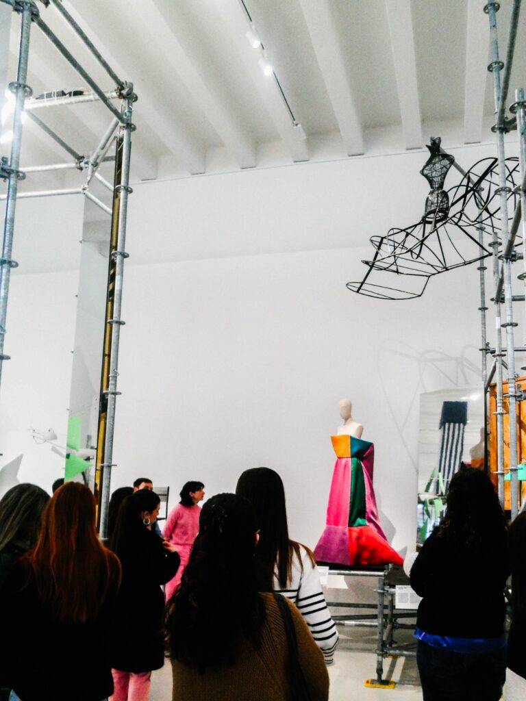 La imagen muestra a un grupo de personas observando una exposición de moda y arte contemporáneo en un museo.