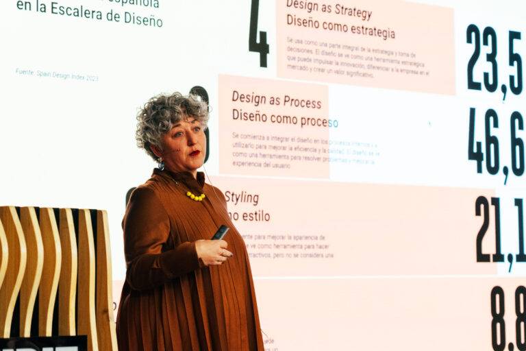 Una mujer con cabello rizado y vestido marrón presenta conceptos de diseño en una conferencia.