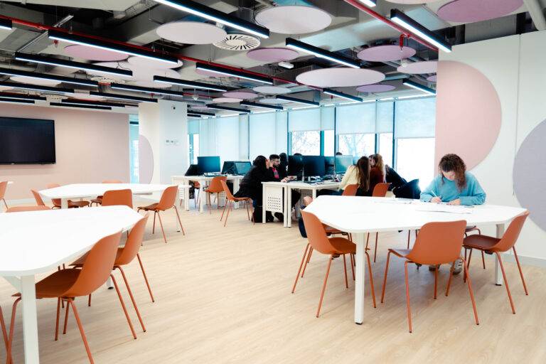 Una moderna sala de estudio con mesas blancas y sillas naranjas, donde varias personas trabajan y estudian.
