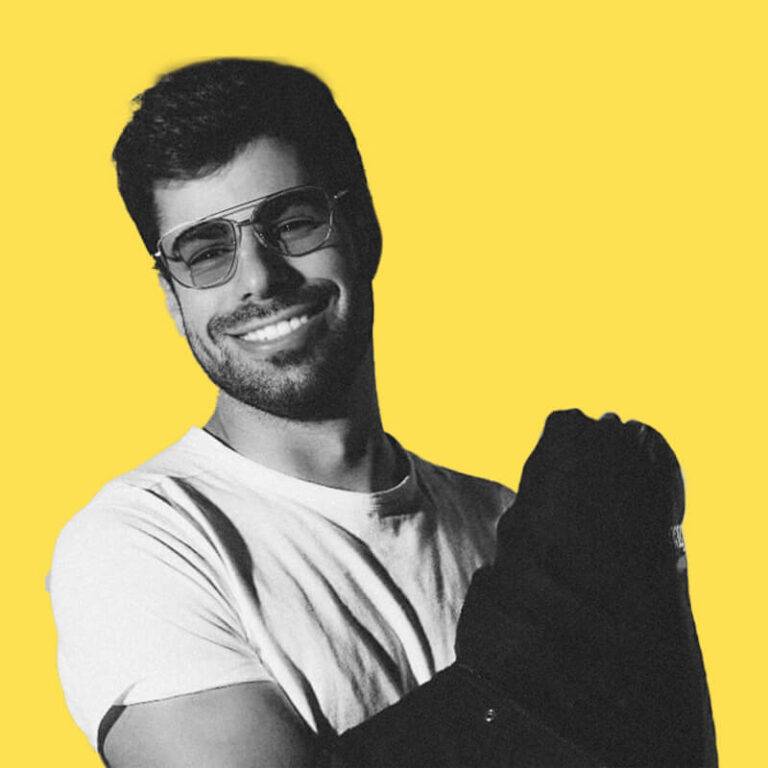 Un hombre sonriente con gafas de sol y una camiseta blanca, posando con un fondo amarillo.