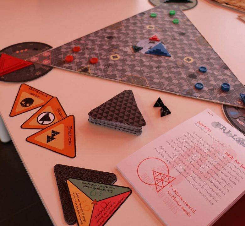 La imagen muestra un juego de mesa con un tablero triangular, fichas de colores y cartas en la mesa.