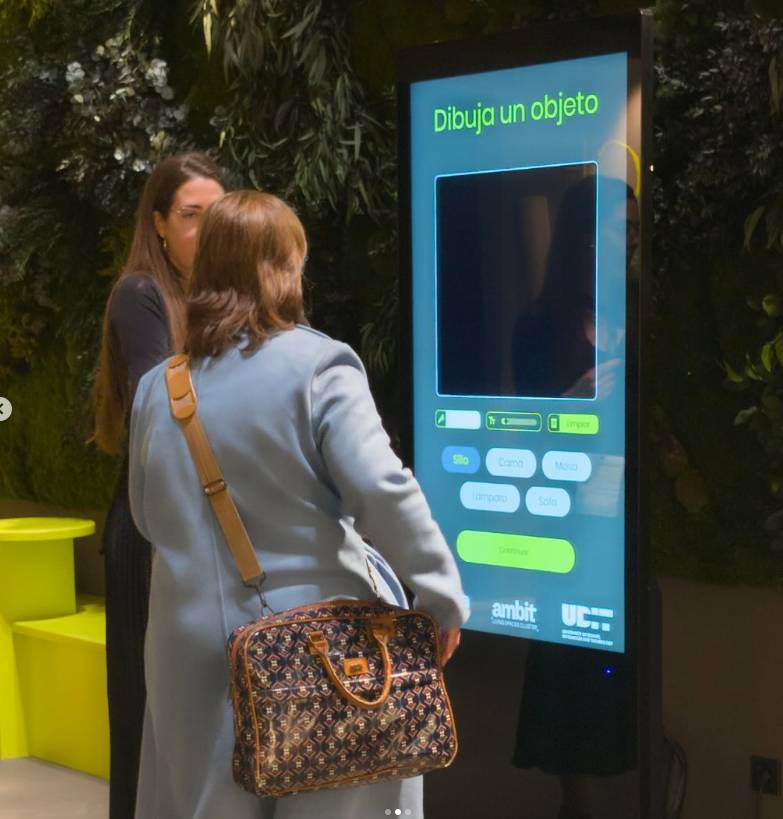 Dos personas interactúan con un kiosco digital que invita a dibujar un objeto.