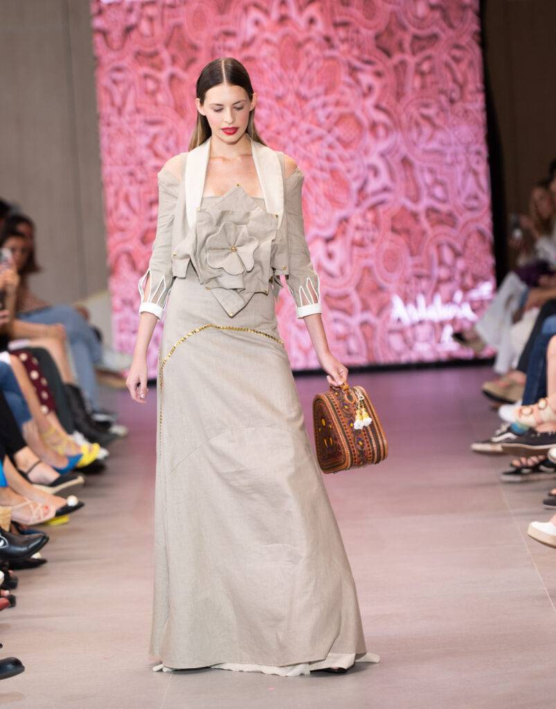 Una modelo desfilando con un vestido elegante y una bolsa destacada en una pasarela.