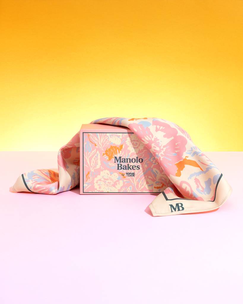 Una caja de Manolo Bakes con un pañuelo de seda decorado sobre un fondo colorido.