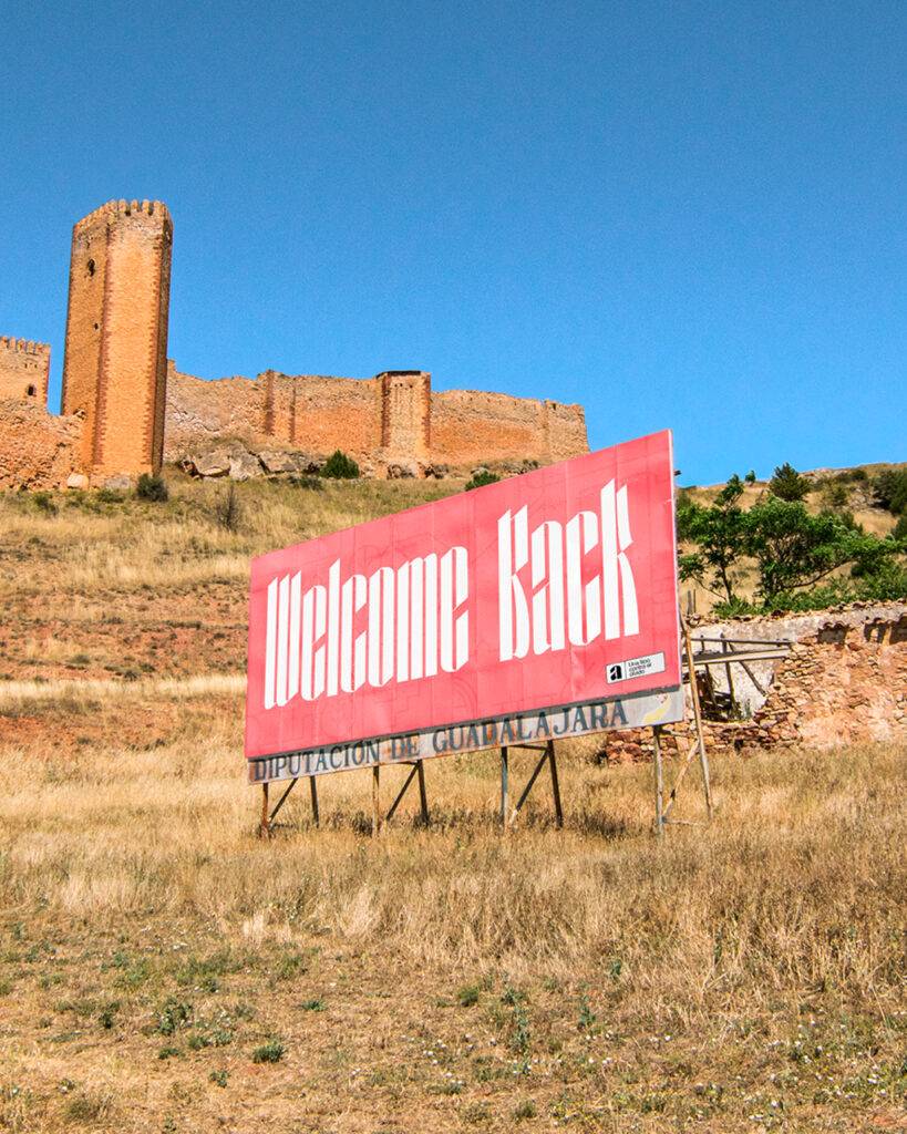 Una pancarta que dice 'Welcome Back' frente a una antigua fortaleza en un paisaje soleado.