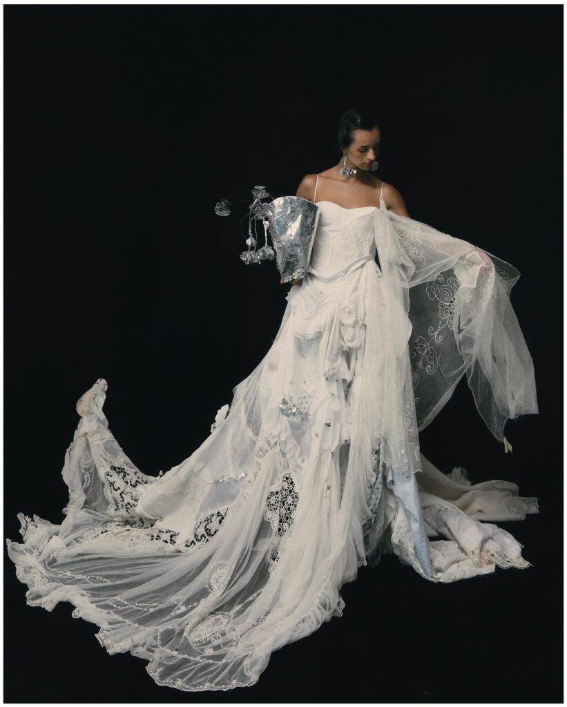Una modelo viste un vestido blanco espectacular con detalles artísticos y un tocado elegante sobre un fondo negro.