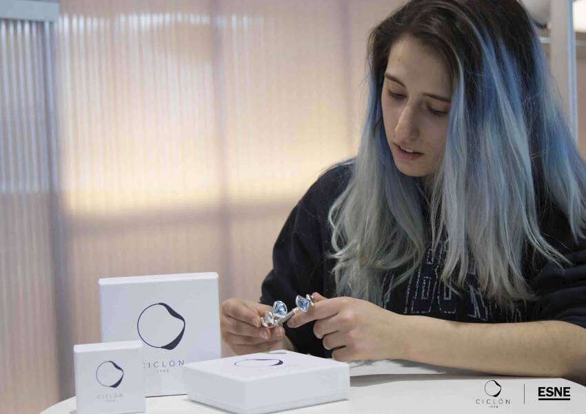Una persona con cabello largo y rubio, observa un objeto plateado mientras lo sostiene con una mano, rodeada de cajas blancas de un producto llamado 'CICLON'.
