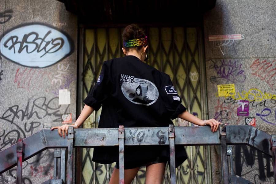 Una persona de espaldas viste una chaqueta negra con un diseño gráfico en un entorno urbano lleno de graffiti.