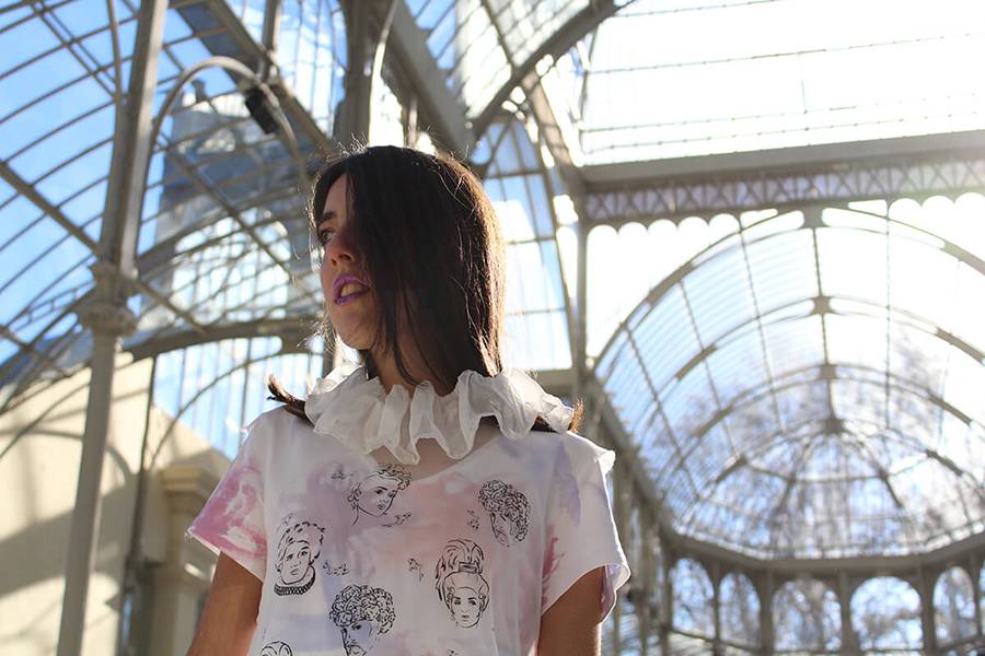Una mujer con una blusa decorada y un collar de volantes, posando bajo una estructura arquitectónica de vidrio.