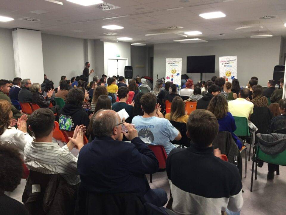 Una multitud de personas sentadas en un salón, escuchando una presentación.