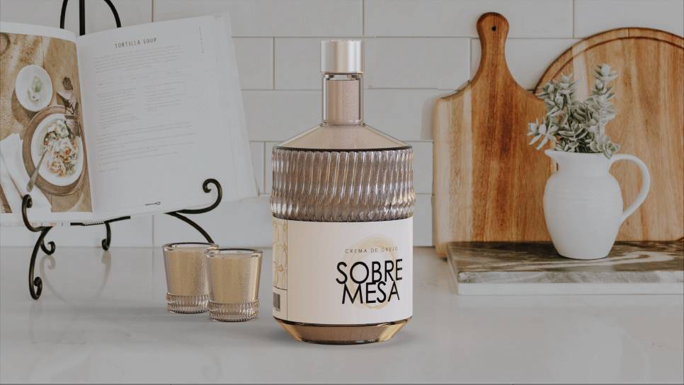 Una botella de licor 'Sobre Mesa' acompañada de dos copas y un libro de recetas sobre una mesa de cocina.