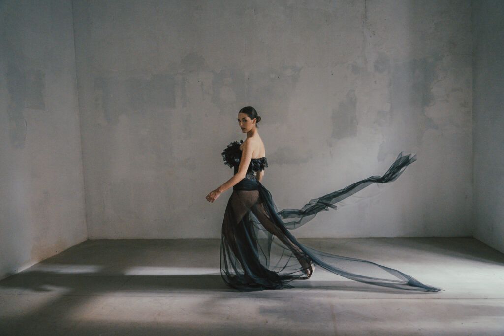 Una modelo camina con un vestido negro que tiene una capa de tela ligera en un espacio minimalista.