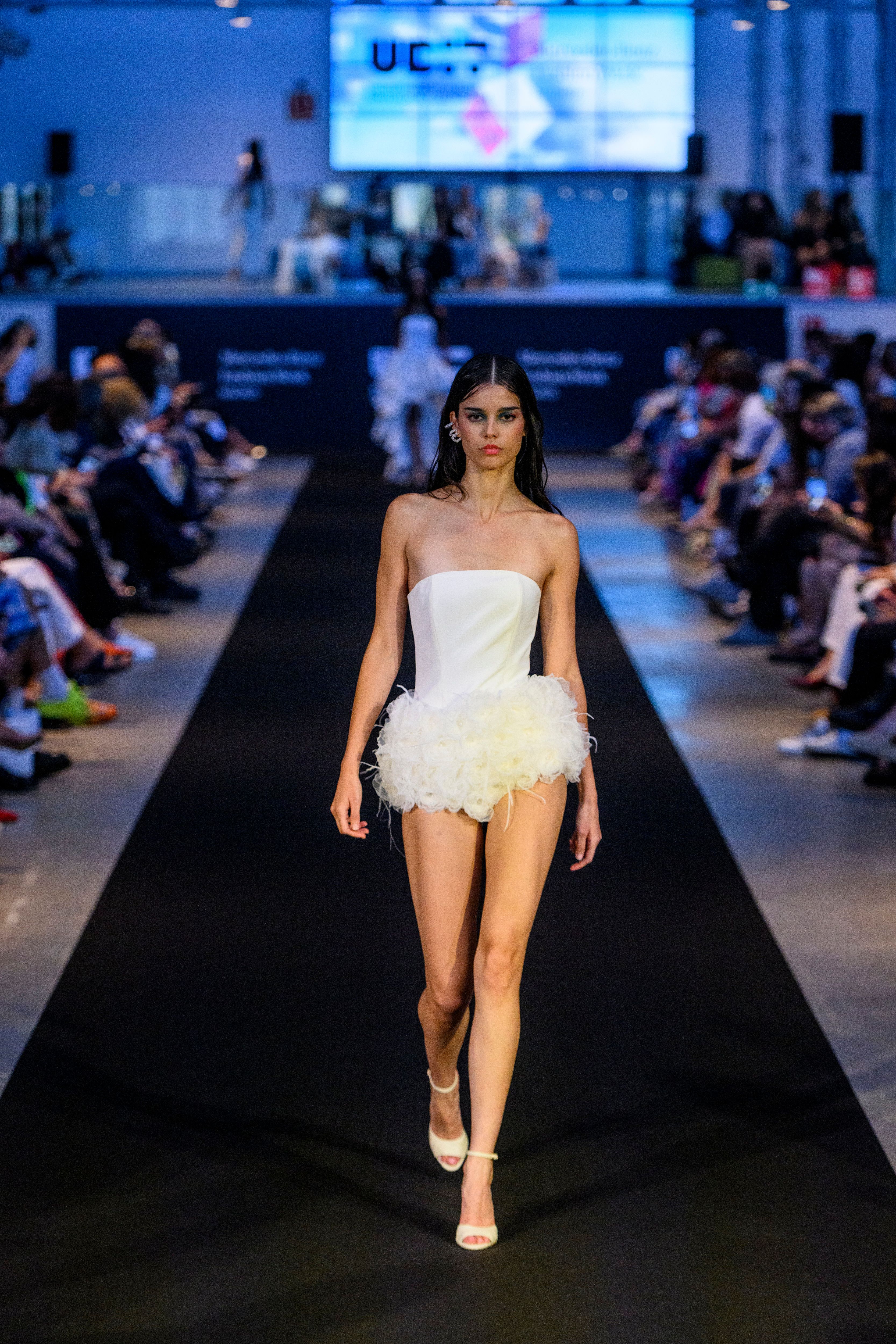 Una modelo camina por la pasarela luciendo un vestido blanco con detalles de plumas en un evento de moda.