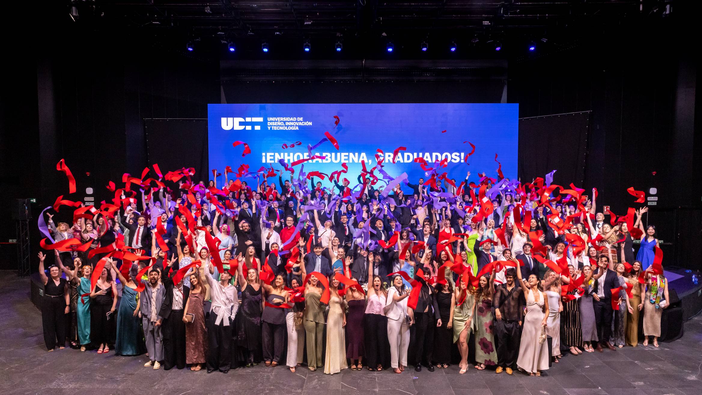Un grupo numeroso de personas celebra su graduación en un evento festivo con cintas rojas en las manos.
