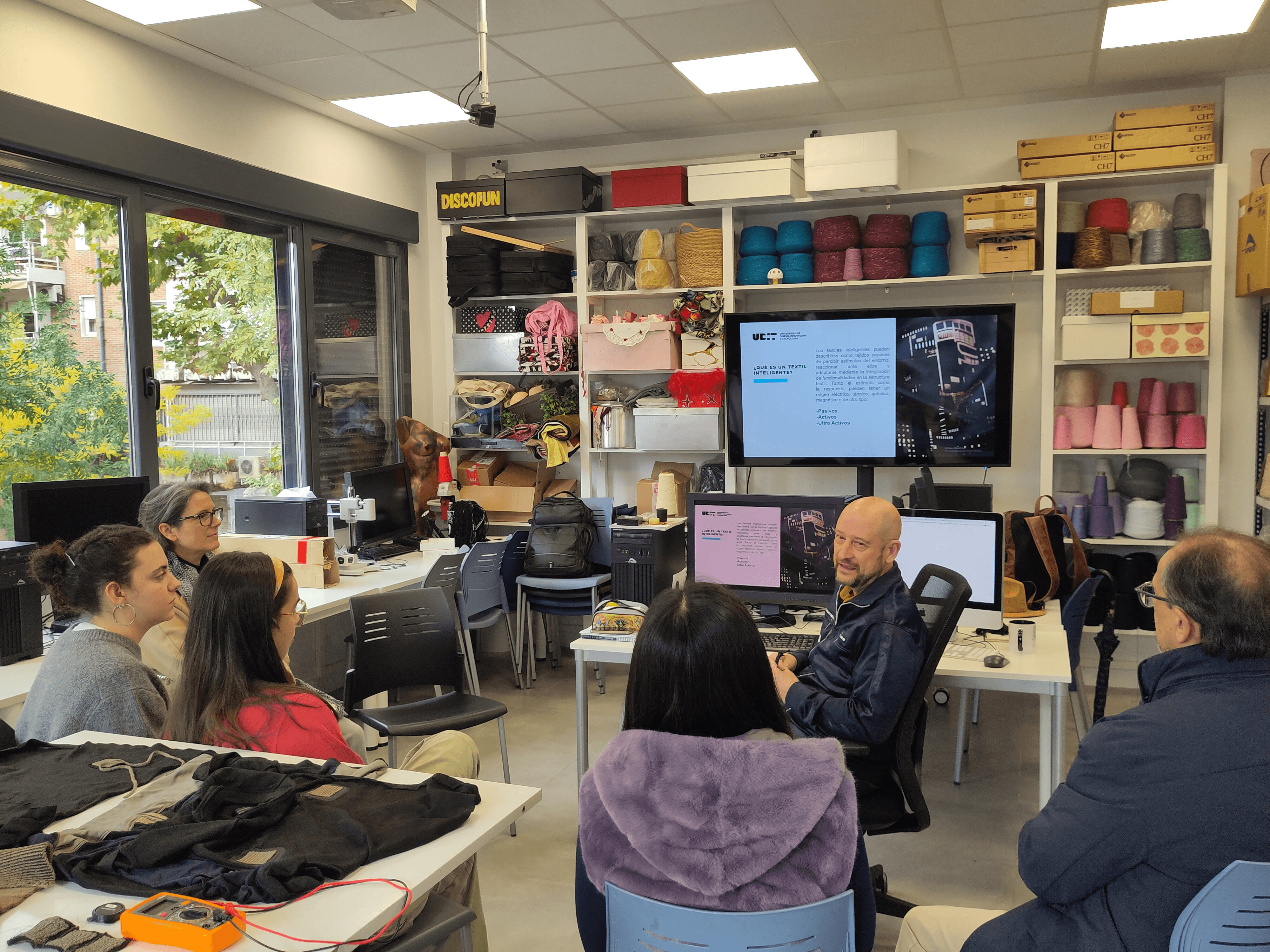 Un grupo de personas se encuentra reunido en un aula con un presentador frente a una pantalla mostrando información.